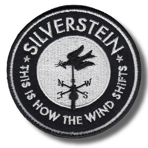 May include: A circular embroidered patch featuring a black and white design. The center displays a weather vane with a bird and cardinal directions. The border text reads SILVERSTEIN and THIS IS HOW THE WIND SHIFTS, separated by two small stars.