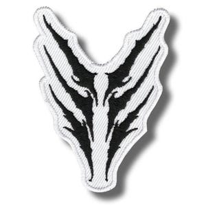 May include: A white embroidered patch featuring a symmetrical black graphic design that resembles claw marks or sharp, jagged streaks. The patch has a distinct V-shaped top and a pointed bottom, with a textured fabric surface and stitched edges.