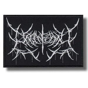 May include: A rectangular black embroidered patch featuring white thread work in a stylized, jagged, and dripping font design. The embroidery creates a dense, web-like pattern of sharp, pointed lines that span the center of the black fabric background.