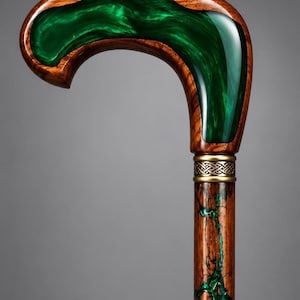 Puede incluir: Un bastón con mango curvo de madera marrón pulida e incrustaciones de resina verde esmeralda con patrones en espiral. Una banda de metal dorado con un intrincado diseño de nudo celta conecta el mango con el cuerpo de madera.