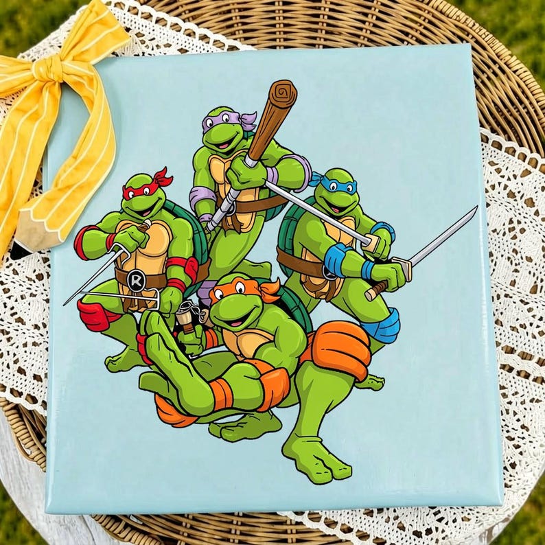 May include: A light blue square canvas featuring a colorful illustration of four green turtles with colored masks, each wielding a different weapon, including a bo staff, katanas, and nunchucks. A yellow striped ribbon is attached to the top corner.