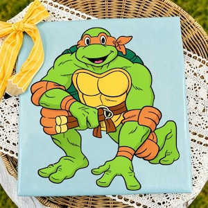 May include: A square canvas print featuring a cartoon illustration of a green Teenage Mutant Ninja Turtle character with an orange mask and arm bands, holding nunchucks, set against a light blue background with a yellow striped pencil bow.