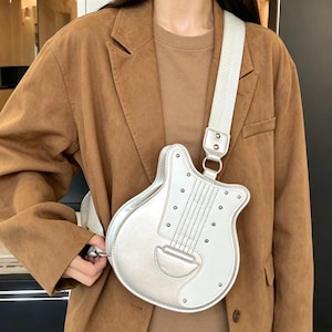 Op de afbeelding: Een zilverkleurige schoudertas in de vorm van een elektrische gitaar, voorzien van een metallic afwerking, decoratieve metalen studs en een brede, verstelbare stoffen schouderband. De tas wordt gedragen over een bruine blazer en bijpassend shirt.