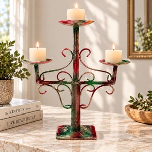 May include: A metal candelabra featuring a central pillar and two side arms with three circular candle plates. The structure is finished in a mottled red and green paint pattern and holds three white pillar candles with lit wicks.