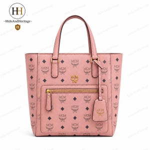 Może przedstawiać: Pudroworóżowa torba typu tote z nadrukiem monogramu MCM w kolorze ciemnobrązowym na całej powierzchni. Torba posiada dwa uchwyty, zewnętrzną kieszeń z przodu ze złotym zamkiem błyskawicznym, złote logo MCM oraz pasującą skórzaną zawieszkę.