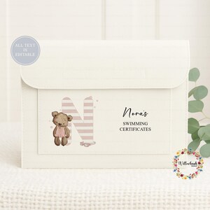 May include: A cream-colored document organizer featuring a large letter N with pink and white horizontal stripes, a brown teddy bear illustration, and the text Nora's SWIMMING CERTIFICATES. A circular badge in the corner states ALL TEXT IS EDITABLE.