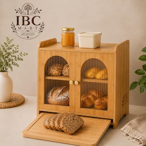 May include: A bamboo bread box with two arched doors featuring fluted glass panels and small gold knobs. The unit has two interior shelves holding various baked breads. A removable bamboo cutting board sits at the base with sliced bread.