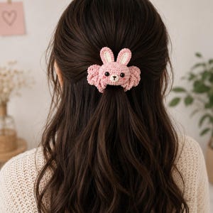 以下が含まれることがあります： ピンク色のクロッシェ編みのヘアアクセサリー。立ち耳、黒いビーズの目、白い口元を持つ小さなウサギの顔がデザインされています。ウサギはヘアクリップに取り付けられており、ダークブラウンの髪の一部をハーフアップにまとめています。