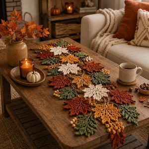 May include: A crochet table runner featuring a repeating pattern of autumn leaves in shades of rust orange, golden yellow, cream, and forest green, accented with small clusters of round berries, laid flat across a wooden coffee table surface.