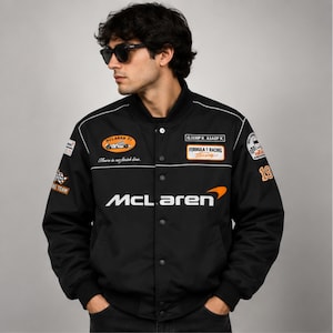 Può includere: Un giubbotto bomber nero con loghi da corsa McLaren Formula 1 in bianco e arancione, inclusi i testi McLaren, Formula 1 Racing e There is no finish line. Il capo presenta una chiusura con bottoni a pressione, polsini a coste e varie toppe.