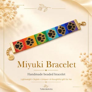 May include: A beaded bracelet featuring a rainbow gradient background of red, orange, yellow, green, blue, and purple beads. Each color section contains a black paw print pattern. The bracelet has gold-toned metal ends, a lobster clasp, and a small round charm.