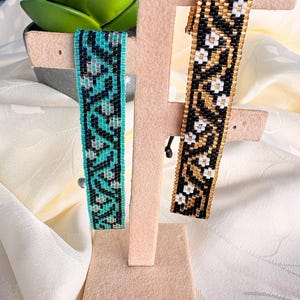 May include: Two woven bead bracelets displayed on a beige stand. The left bracelet features a teal, black, and silver geometric pattern. The right bracelet features a gold, black, and white geometric pattern. Both are crafted from small beads.