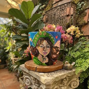 May include: A hand-painted ceramic plant pot featuring a portrait of a person with long brown hair, a green garment, and a green halo-like design against a blue background with small red hearts, resting on a circular wooden base.