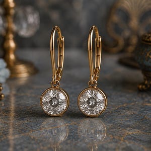 May include: A pair of gold leverback earrings featuring round, clear, faceted stones set in a circular gold bezel. The earrings have a polished gold finish and a classic drop design, with the stones reflecting light from their multiple facets.