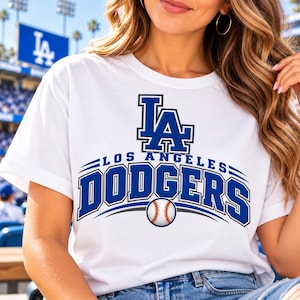 Pode incluir: Uma t-shirt branca de manga curta com uma impressão gráfica azul do logótipo LA acima do texto LOS ANGELES DODGERS numa fonte azul a negrito, com uma ilustração de uma bola de basebol posicionada abaixo do nome da equipa na frente.