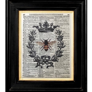 May include: Framed vintage dictionary page art print featuring a bee design. The bee is centered within a crown, laurel wreath, and ribbon design. The print is in a black frame with a cream-colored mat.