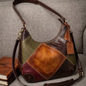May include: A hobo-style shoulder bag featuring a patchwork design of dark brown crocodile-embossed leather, olive green woven texture, and a smooth tan centre panel. The bag includes a brown leather tag with a tree logo and the text HERENCIA.
