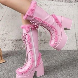 May include: A pair of knee-high pink boots featuring a thick platform sole and a chunky block heel. The boots are accented with pink faux fur trim along the seams and collar, and include a front lace-up closure with matching pink laces and a side zipper.