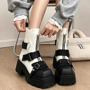 May include: White ankle boots featuring a black square toe, a thick black lug sole, and a front silver zipper closure. The boots are accented with black adjustable straps and buckles across the front and sides, providing a secure fit and a distinct look.