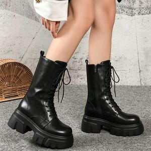 May include: A pair of black mid-calf boots featuring a thick, chunky lug sole, a lace-up front closure with matching black laces, and a vertical side zipper on the inner shaft. The boots have a smooth, matte finish and a rounded toe design.