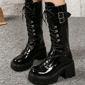 May include: A pair of black patent leather mid-calf boots featuring a thick lug sole, a chunky block heel, and a front lace-up closure. The boots include silver-toned metal eyelets and a decorative strap with a silver buckle at the top shaft.