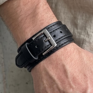 Op de afbeelding: Een zwarte leren manchetarmband met een zilverkleurige metalen gespsluiting. De armband heeft een gladde textuur met zichtbare stiksels langs de randen en een riemontwerp dat om de pols wikkelt, vastgezet met een metalen gesp met één doorn.