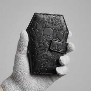 May include: A black coffin-shaped wallet featuring an embossed pattern of skulls, skeletons, bats, and spiders. The wallet has a secure snap-button closure flap on the side and a smooth, matte finish across the entire exterior surface.