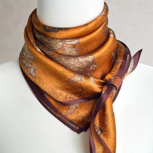 Könnte beinhalten: Ein quadratisches Tuch in sattem Bernstein-Orange mit einem metallisch goldenen Blumenmuster und einem dunkelbraunen Rand. Das Tuch wird mit einem kleinen, runden braunen Knopfverschluss gehalten. Der Text REAL PRODUCT PHOTOS ist unten sichtbar.