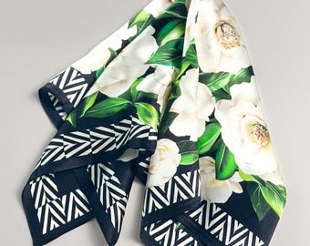 May include: A square scarf featuring a large-scale floral print of white camellia blossoms and green leaves set against a black background. The edges of the scarf are finished with a bold black and white geometric chevron border pattern.