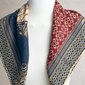 Op de afbeelding: Een vierkante sjaal met een meerdelig patroon in donkerrood, marineblauw en crème. Het ontwerp bevat herhalende goudkleurige kettingschakels, stijgbeugels in ruiterstijl en kwastmotieven, afgewerkt met een smalle, crèmekleurige rand.