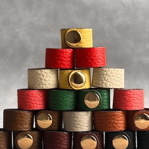 Op de afbeelding: Een piramidevormige stapel van gestructureerde leren armbanden in diverse kleuren zoals geel, rood, crème, groen, bruin en zwart. Elke armband is voorzien van een gepolijste goudkleurige, ronde drukknoopsluiting aan de voorzijde.