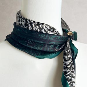 May include: A square scarf featuring a dark green and black paisley pattern on one side and a black and white geometric Greek key border on the other. The scarf is secured with a small, round, metallic gold-toned button fastener at the neck.