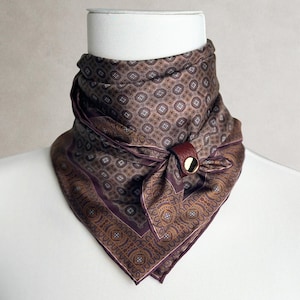 May include: A brown and purple patterned silk neckerchief featuring a geometric and paisley design, secured with a dark red leather ring featuring a gold-tone metal stud. The fabric displays a repeating square motif with small white accents.