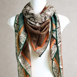 Pode incluir: Um lenço quadrado com um padrão paisley complexo em tons de castanho escuro, creme e verde suave. O lenço tem uma faixa laranja distinta na borda e uma bainha verde escura, drapeado em torno de um manequim branco.