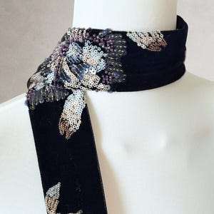 May include: A black velvet choker scarf featuring intricate floral embroidery crafted from shimmering silver, purple, and bronze sequins. The design includes a dense cluster of sequins on one side and smaller scattered floral motifs along the band.