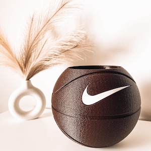 Puede incluir: Un recipiente esférico de color marrón oscuro con una superficie texturizada que recuerda a un balón de baloncesto, con un prominente logotipo Nike swoosh blanco en la parte frontal. El objeto tiene una abertura hueca en la parte superior.