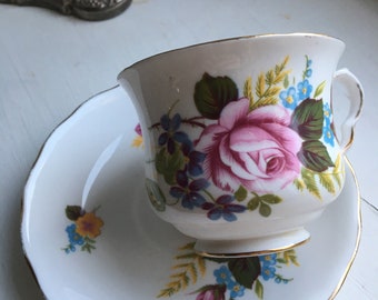 Può includere: Una tazza da tè e un piattino bianchi con un motivo floreale. La tazza e il piattino hanno un bordo dorato. Il motivo floreale presenta rose rosa, nontiscordardime blu e foglie verdi.