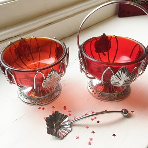 May include: Two red glass bowls with silver leaf accents and silver wire frames. One bowl has a handle and the other does not. A silver spoon with a flower-shaped head is in the foreground.