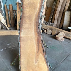 Pode incluir: Uma placa de madeira vertical de borda natural com uma superfície de grão liso em tom castanho claro, apresentando bordas naturais, ásperas e cobertas de casca em ambos os lados. A placa exibe uma variação de cor entre o centro pálido e as bordas.
