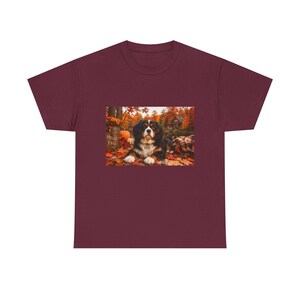 May include: A maroon short-sleeve t-shirt featuring a graphic print of a Bernese Mountain Dog lying on a plaid blanket amidst autumn leaves. The scene includes a pumpkin, a bucket with the text HELLO FALL, and a house in the background.