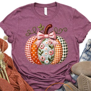 May include: A heather maroon short-sleeve t-shirt featuring a central graphic of a pumpkin with segments in orange gingham, white polka dots, and green plaid, decorated with a pink bow and floral embroidery, alongside a rust-colored knit sweater.