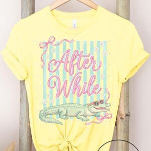 May include: Yellow t-shirt with the text "After While" in pink script, a green alligator graphic, and pink and blue accents. The shirt is hanging on a wooden rack.