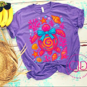 May include: A purple t-shirt with a colorful turtle design, surrounded by coral, seashells, and starfish. The turtle has a blue bow and a spiral shell. A straw hat and bananas with sunglasses are next to the shirt.