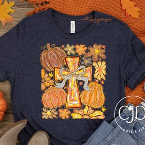 May include: Navy blue t-shirt with a fall-themed graphic featuring pumpkins, flowers, and a cross. The image includes a pair of gray ankle boots, blue jeans, and autumn leaves. A pumpkin and a small white gourd are also visible.