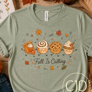 May include: A sage green short-sleeve t-shirt featuring a graphic of four smiling baked goods, including a pie slice, cinnamon roll, tart, and cupcake, with the text "Fall Is Calling" surrounded by small autumn leaves and stars.