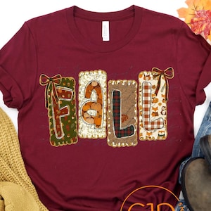 May include: A maroon short-sleeved t-shirt featuring the word FALL in large, decorative block letters. Each letter has a unique pattern including pumpkins, plaid, and woodland animals. The shirt is displayed with a yellow knit sweater and blue jeans.