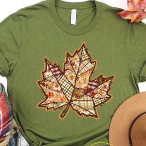 May include: Olive green t-shirt featuring a large, patchwork-style maple leaf design in autumn colors. The leaf design incorporates various plaid and floral patterns. A brown hat and a plaid scarf are also visible.