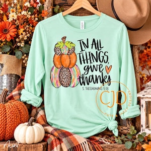 May include: A light mint green long-sleeve t-shirt featuring a graphic of stacked pumpkins with leopard print and colorful stripes. The text reads In all things, give thanks and 1 Thessalonians 5:18 in black script lettering.