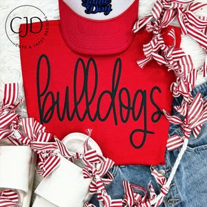 May include: A red sweatshirt with the word "bulldogs" in black script. A red and white trucker hat with the text "Game Day" is above the sweatshirt. White sandals and red and white striped ribbons are also in the image.
