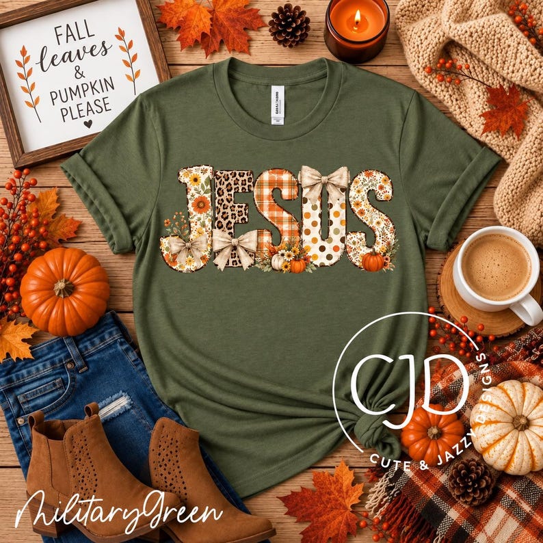 May include: An olive green short-sleeve t-shirt featuring the word JESUS in large letters. Each letter has a unique pattern including sunflowers, leopard print, orange plaid, and polka dots, accented with small bows and pumpkin illustrations.